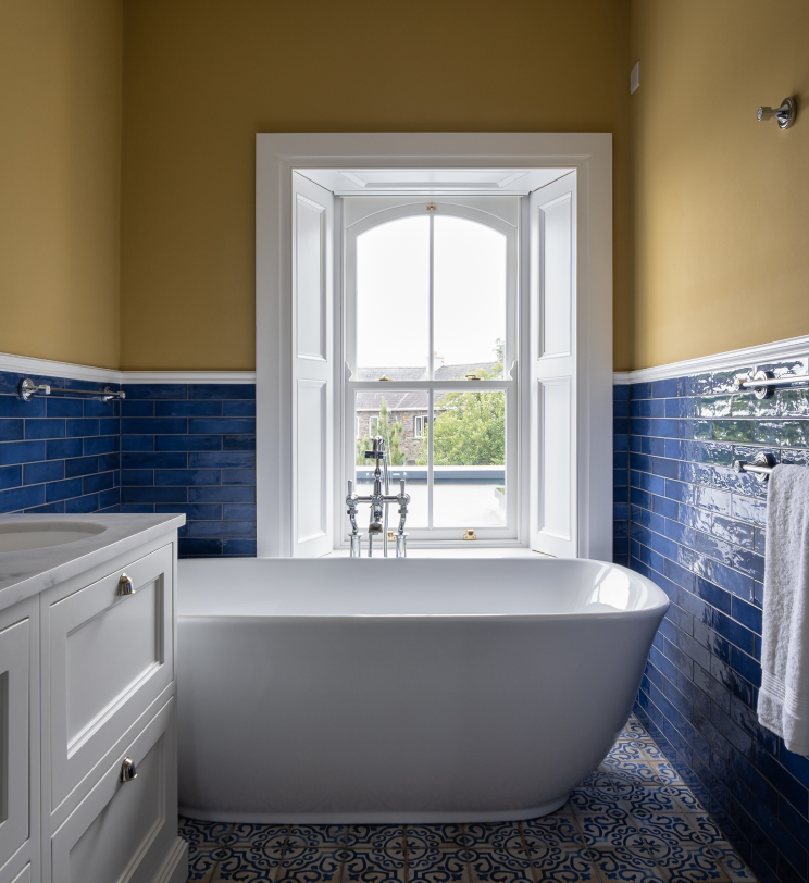 A white bathtub below a window in a bathroom with blue tiles and yellow walls.