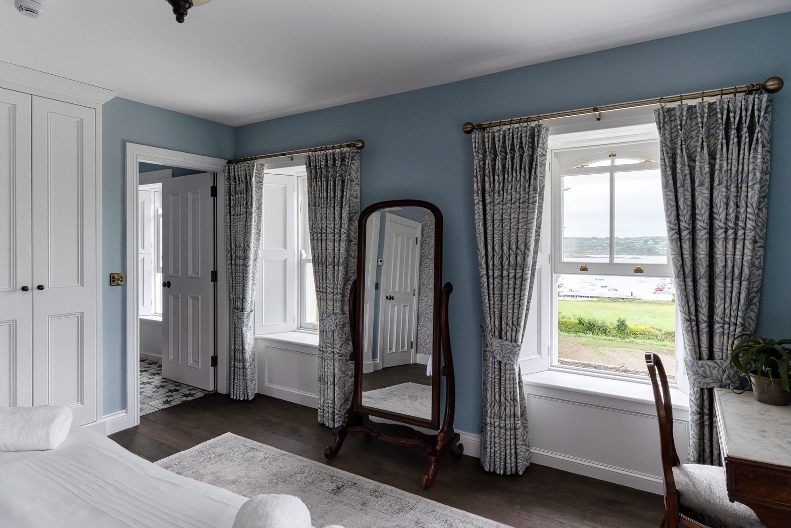 A blue bedroom with a mirror and two windows.