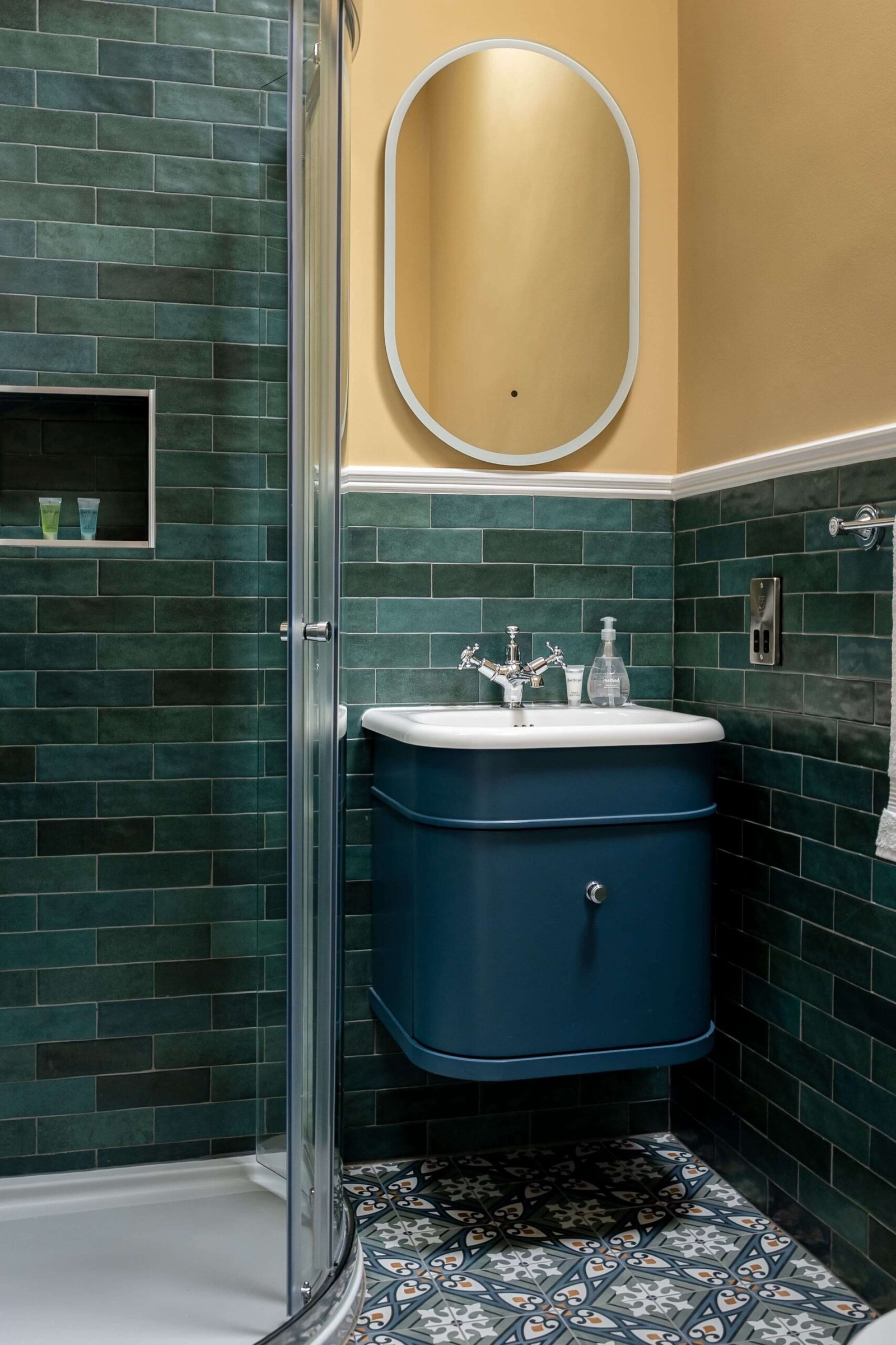 A bathroom with green tiles, a glass shower, a blue vanity sink, and patterned floor tiles.