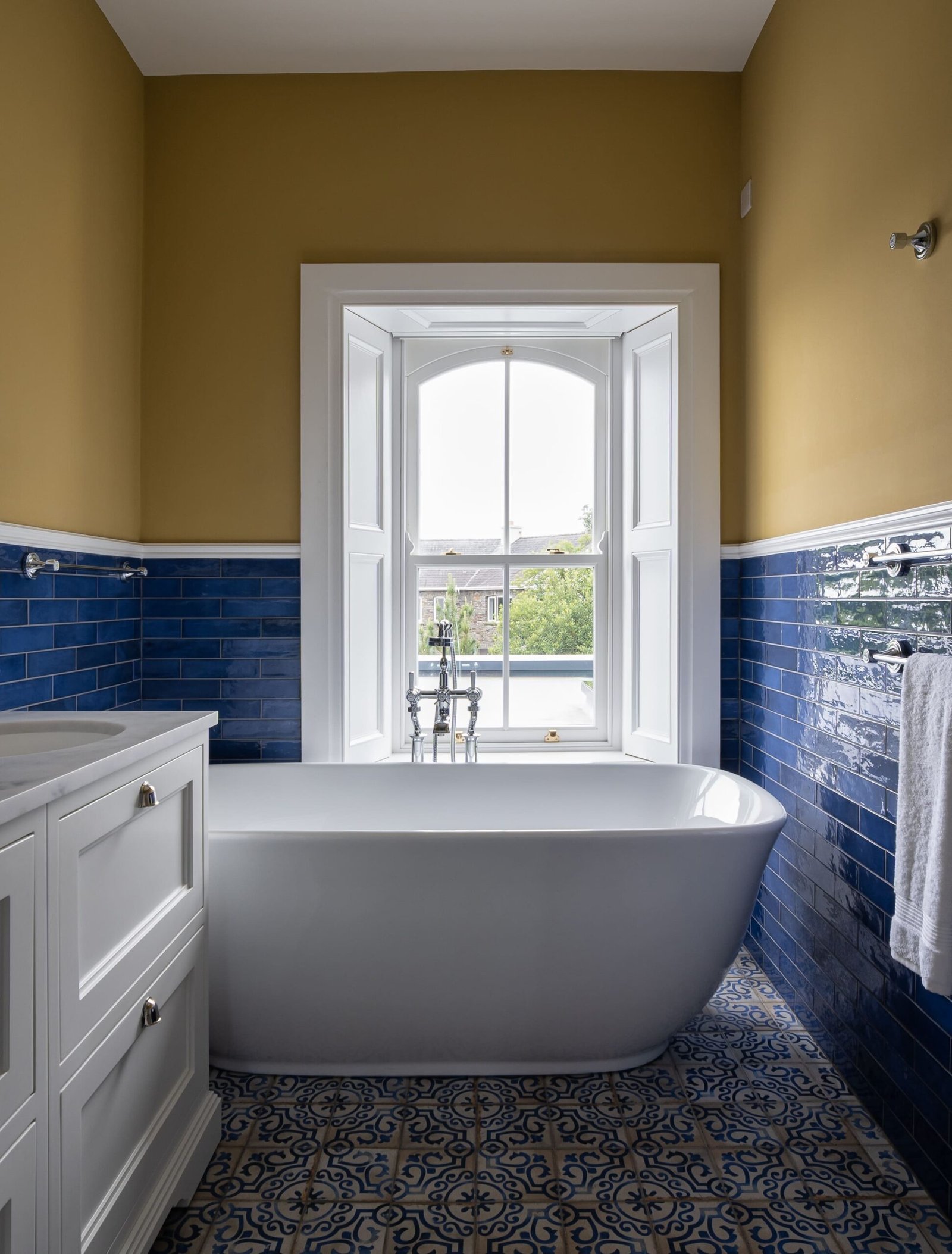 A white bathtub below a window in a bathroom with blue tiles and yellow walls.