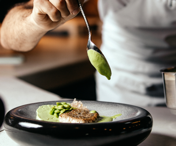 A chef carefully plating a dish by adding a green sauce over a piece of fish with a spoon.