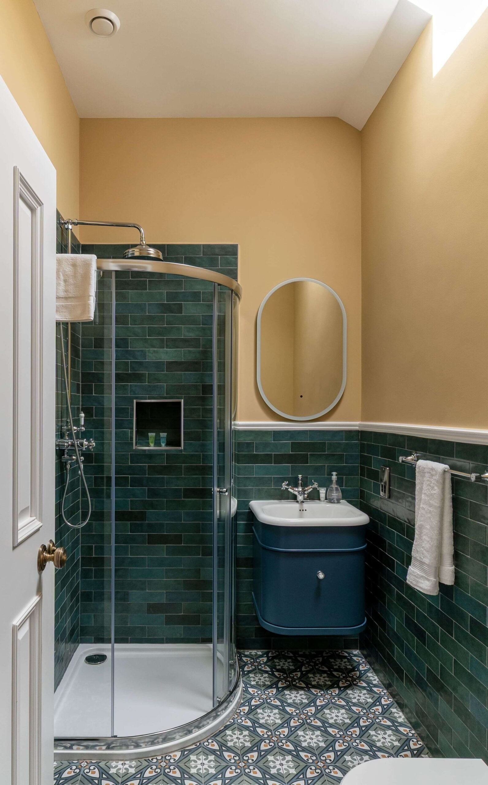 A bathroom with green tiles, a glass shower, a blue vanity sink, and patterned floor tiles.