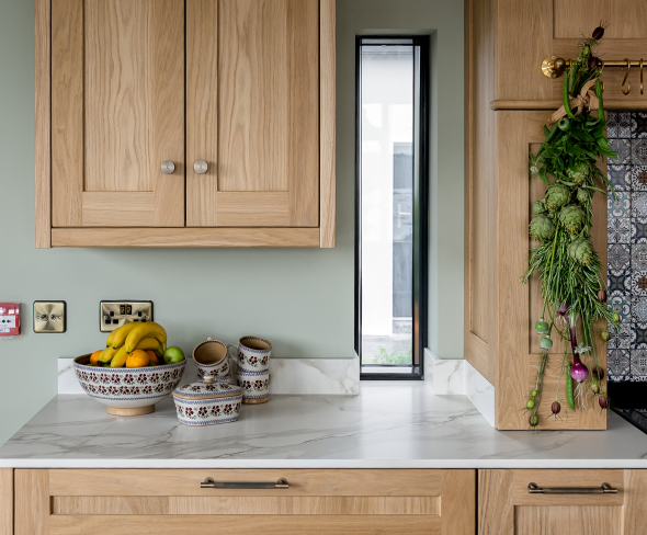 A marble kitchen counter with wooden cabinets, a patterned bowl of bananas and oranges, and a narrow vertical window.