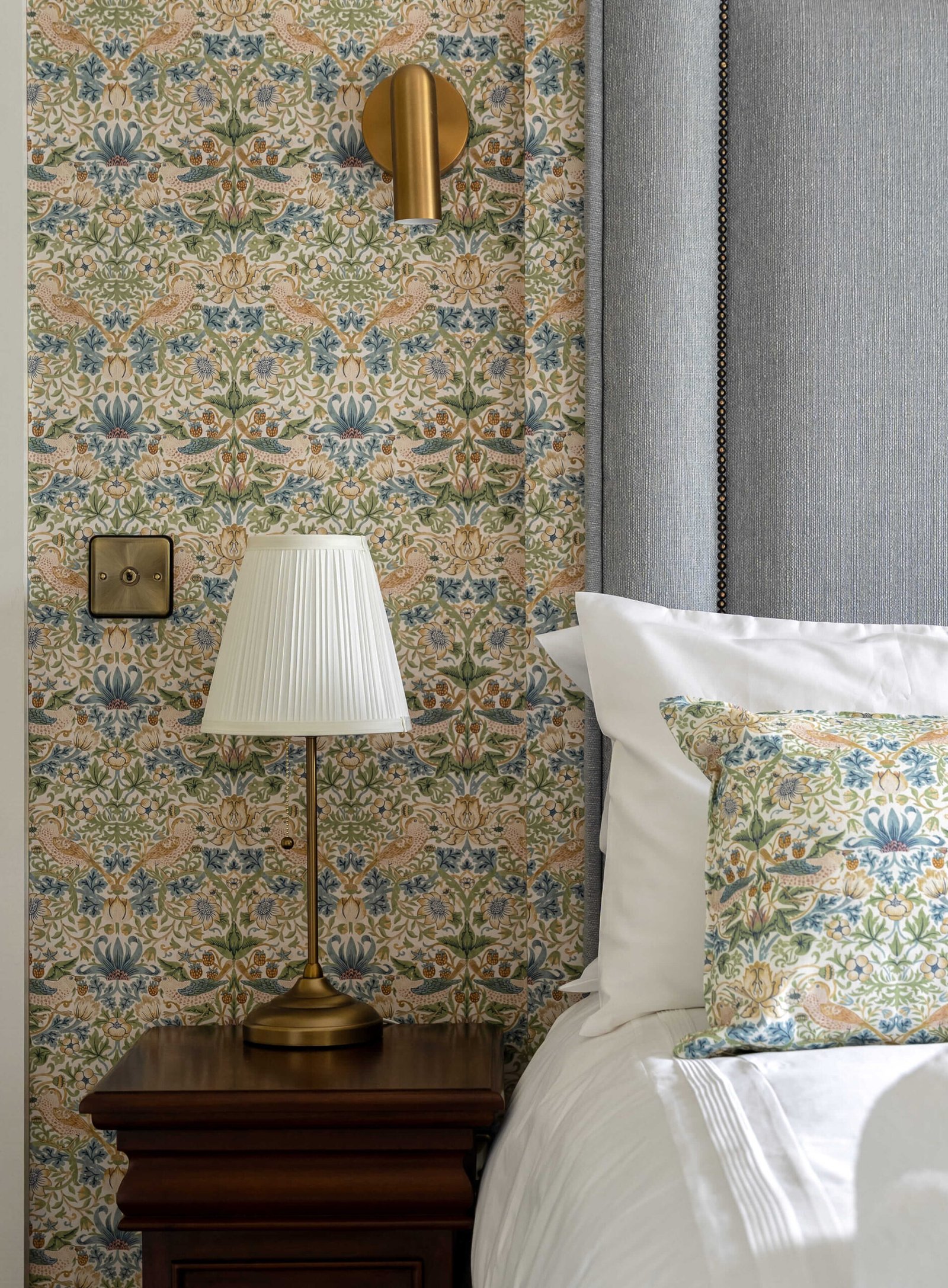 A bedside table with a lamp against a patterned wall next to a grey headboard.