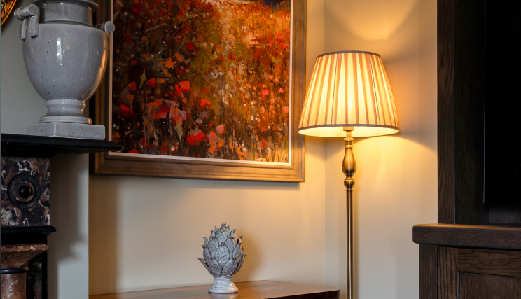 A lit floor lamp standing next to a framed painting of a red flower field, with a small decorative statue on a wooden table below.