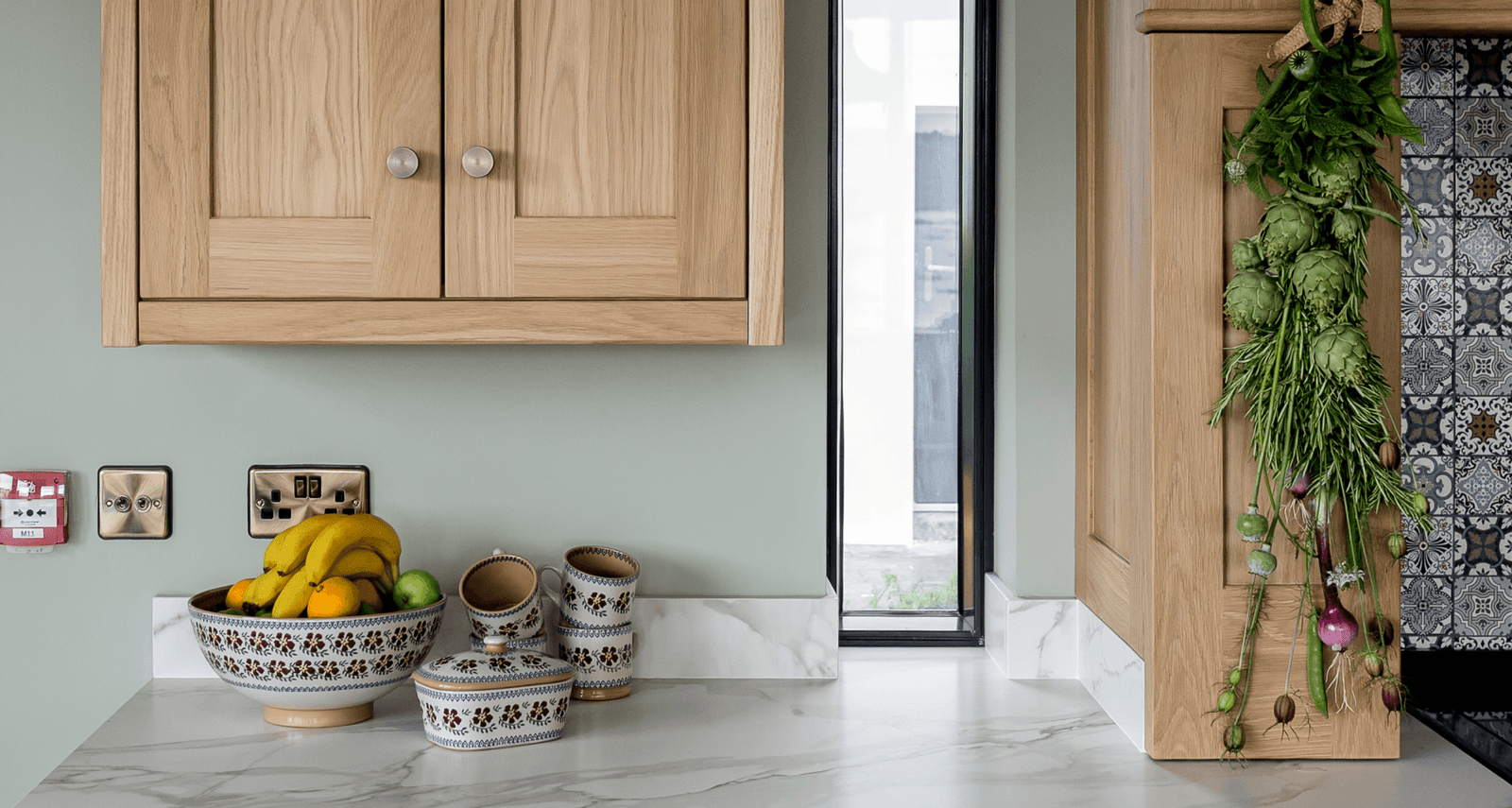 A kitchen counter with light wood cabinets, a patterned bowl of fruit, and hanging herbs.
